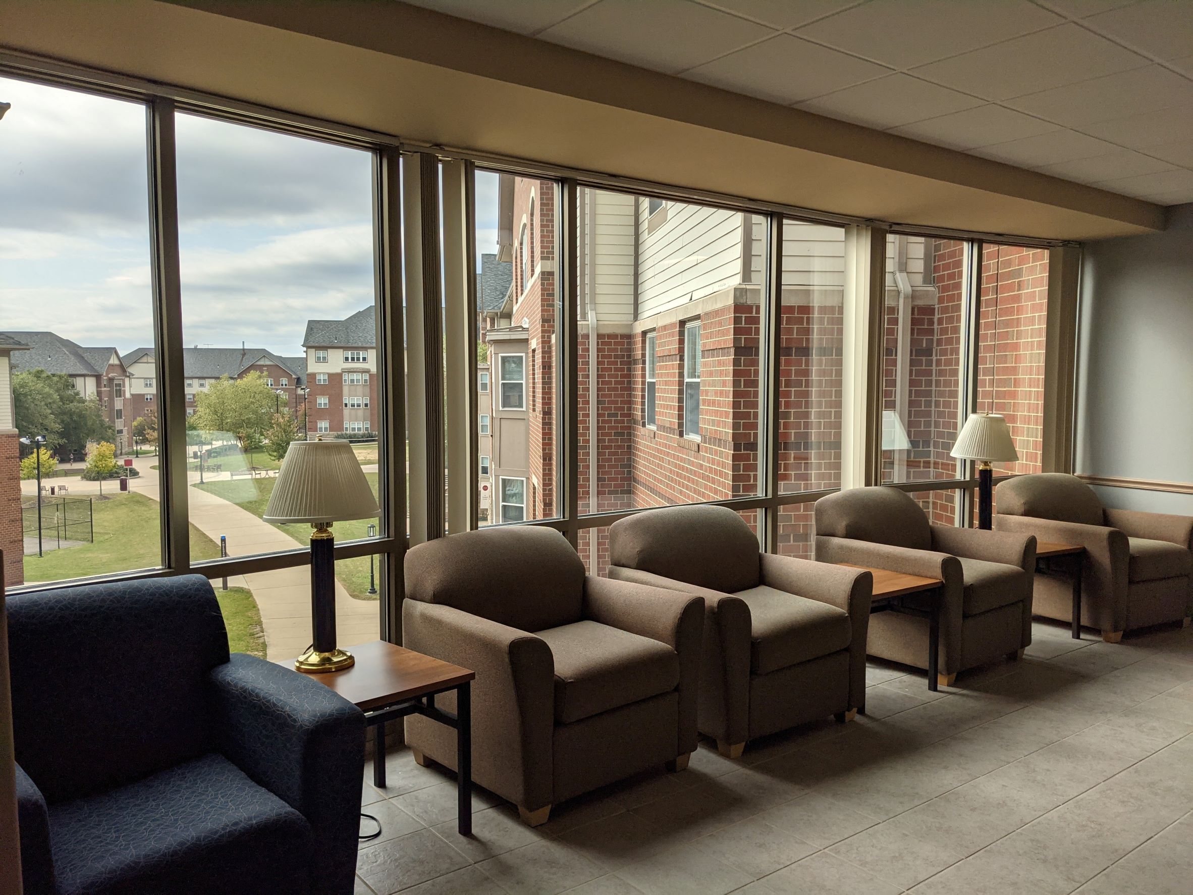Image of a dormitory lounge with chairs along a large window space overlooking a campus courtyard with traditional red brick buildings in the background