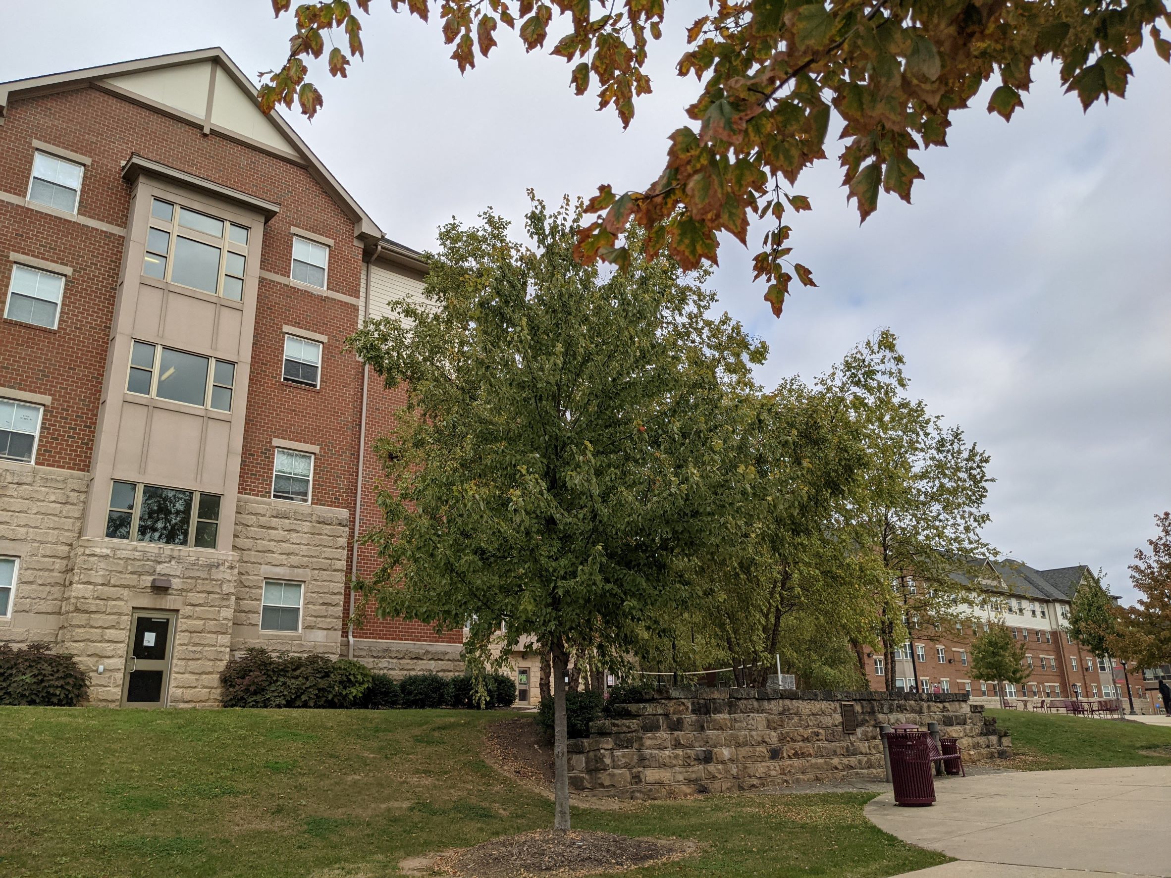 Large red brick gable with tan accents in contemporary living learning student housing community dormitory building with stone retaining wall behind plaza in foreground