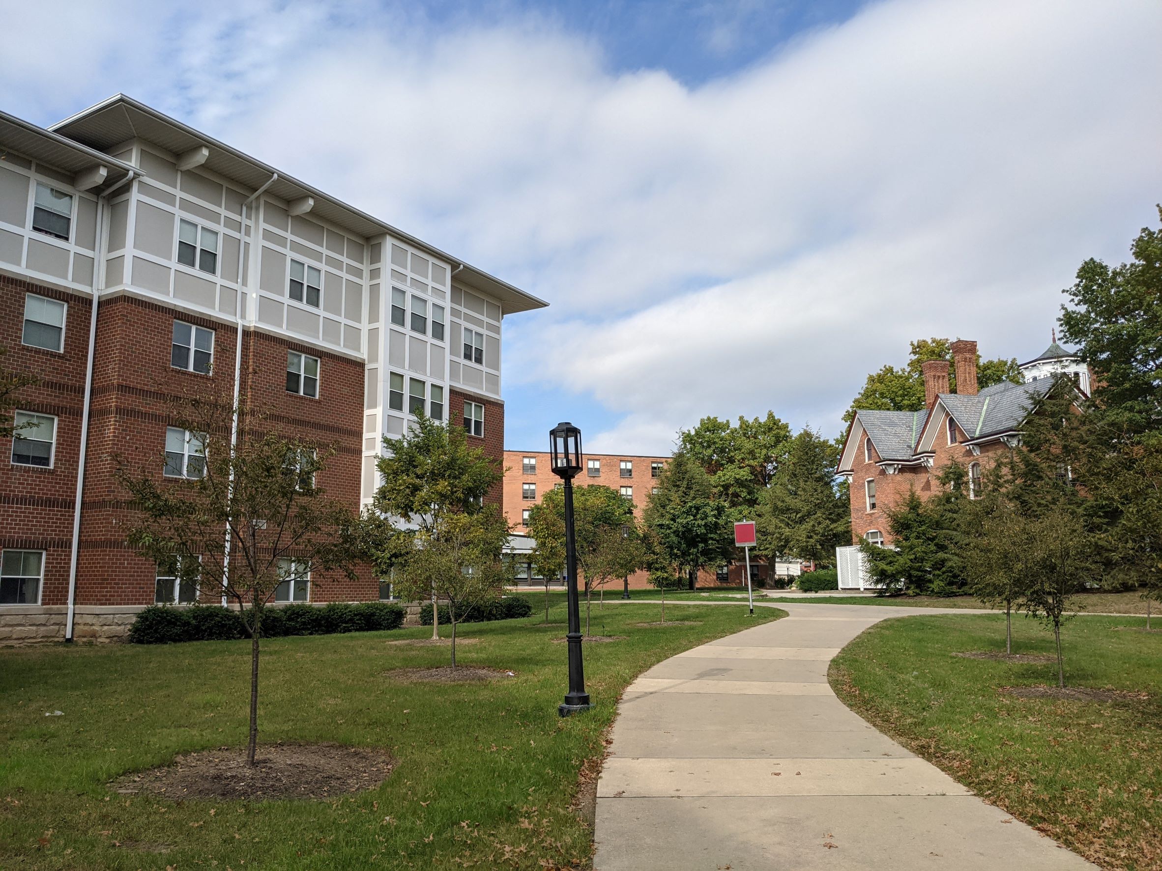 Arts & Crafts style living learning student housing community dormitory building with pink brick and white Tudor style board and batten clerestory and bay window, with large overhangs, sidewalk in foreground with campus alumni house of similar architectural style in background