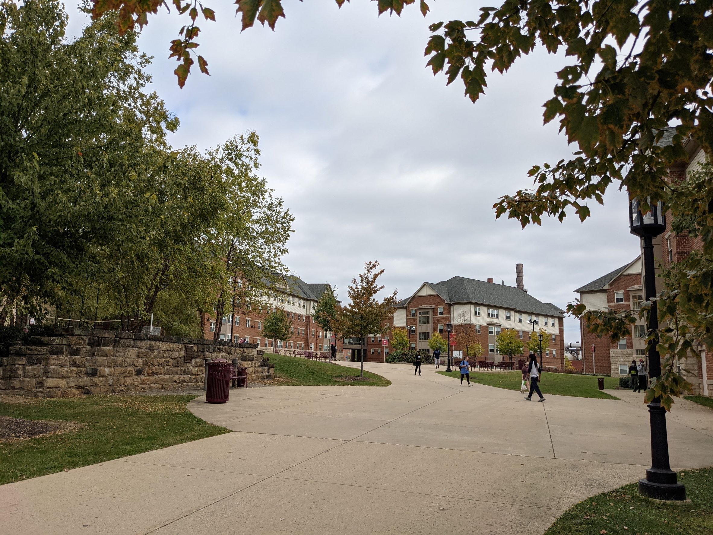 Large plaza area with students walking around with old stone retaining wall and plaque in foreground, traditional red brick living learning student housing community dormitory buildings in background