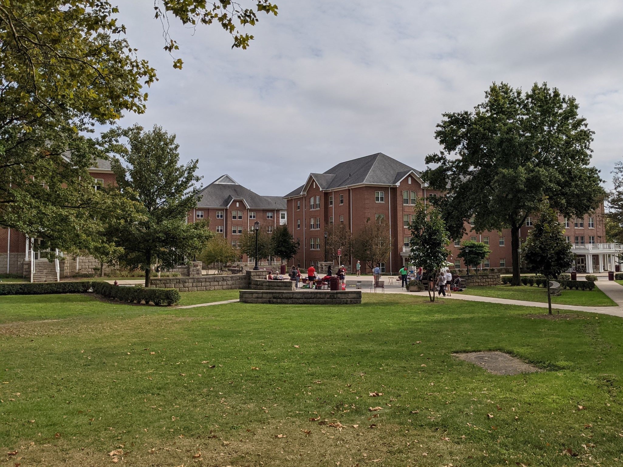 Large campus green with students gathered in plaza with Historical looking pink brick residence hall with many gabled roofs, white front porch and trim, truncated dormer windows and brick quoining, very classical looking student housing building in background