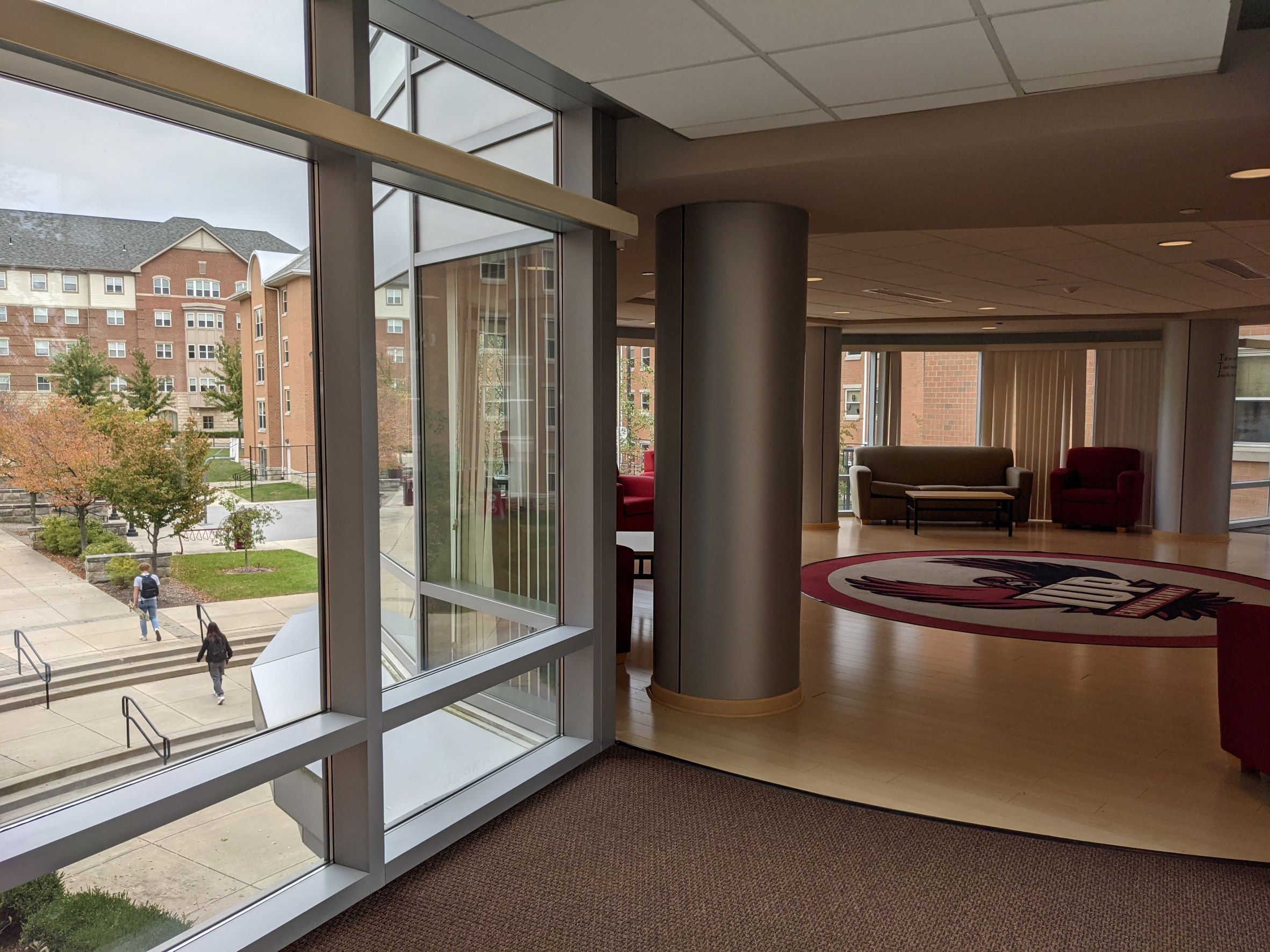 Large community lounge space with IUP school logo on carpet inlaid into hardwood floors, round metal columns with glass everywhere overlooking outdoor courtyard and plaza beyond with  living learning student housing community dormitory building in the background