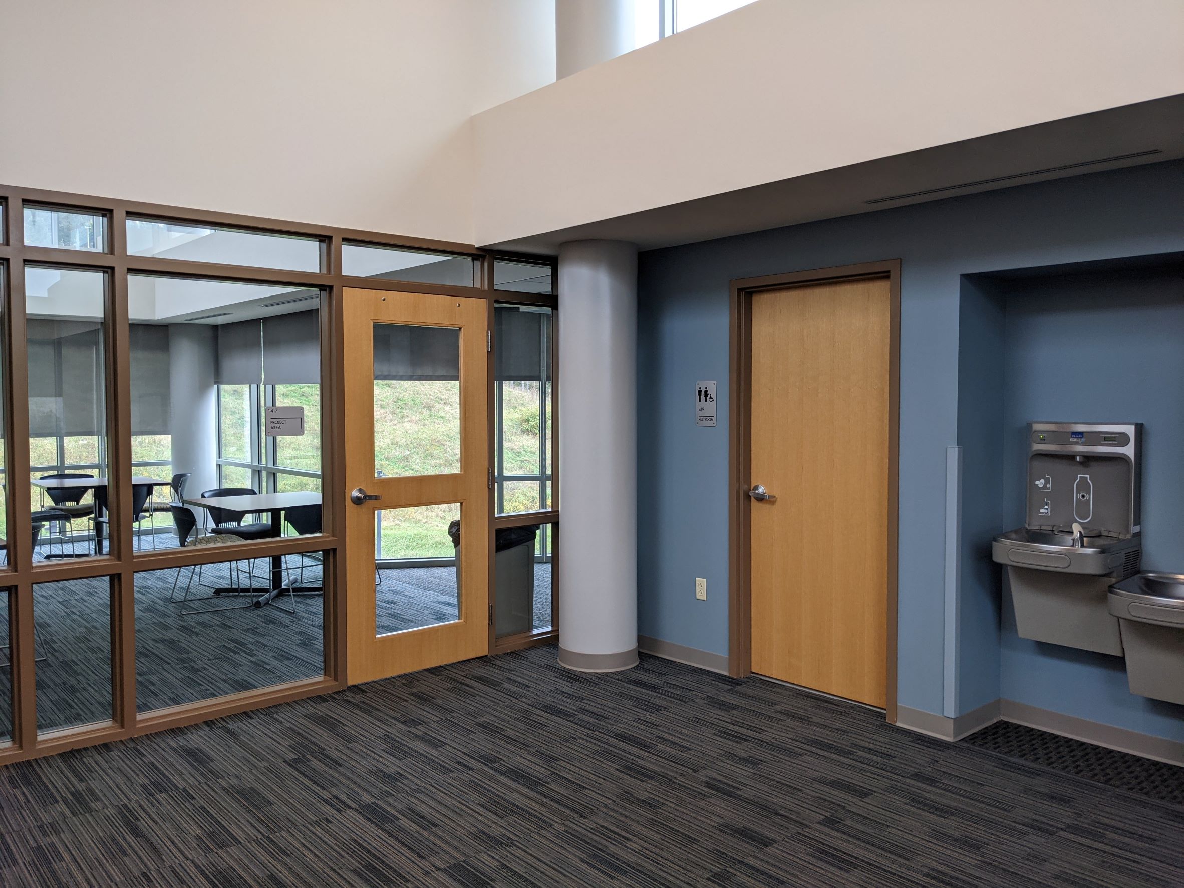 Lobby area of dormitory with cloud blue accent wall, with white bulkheads above, natural wood stained honey colored doors, and glass windows into adjacent lounge space with tables and chairs visible through windows, and wooded trees/hillside beyond, water cooler/drinking fountain with modern paint swatch  looking carpet in foreground