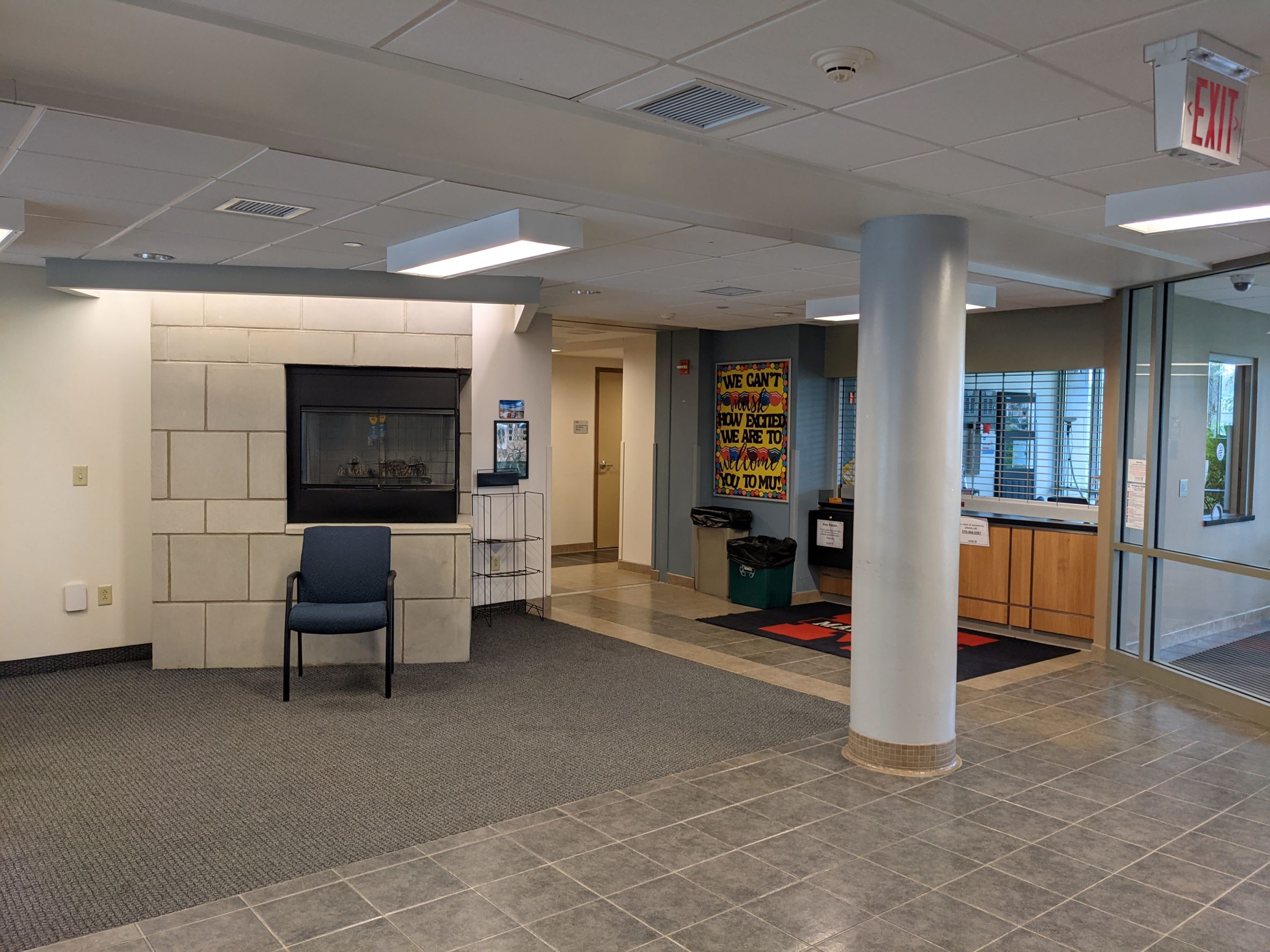 View of residence hall student housing building lobby with reception desk beyond, ceramic floor tile with carpet inlay area around stone corner style (2 sided) fireplace and doors into residents' wings beyond