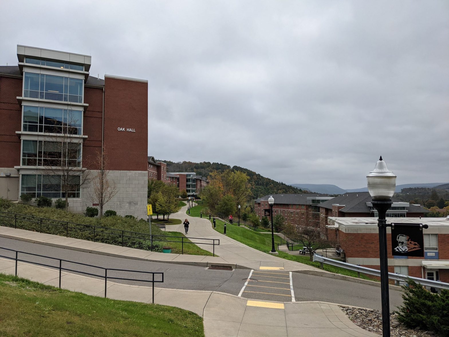 Glass Box projecting from contemporary red brick with stone base living learning student housing community dormitory building overlooking sidewalk above steep hillside with another red brick dormitory building below, mountains in the background