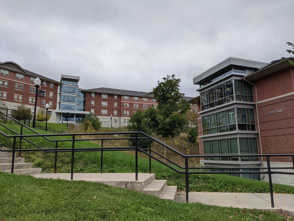 Contemporary large glass box jutting out from contemporary red brick living learning student housing community dormitory building and second red brick with stone base dormitory beyond with another glass box inserted into middle of building up on a hill, with stairs and grass in the foreground. Fraternity and sorority letters can be seen in the closest windows.