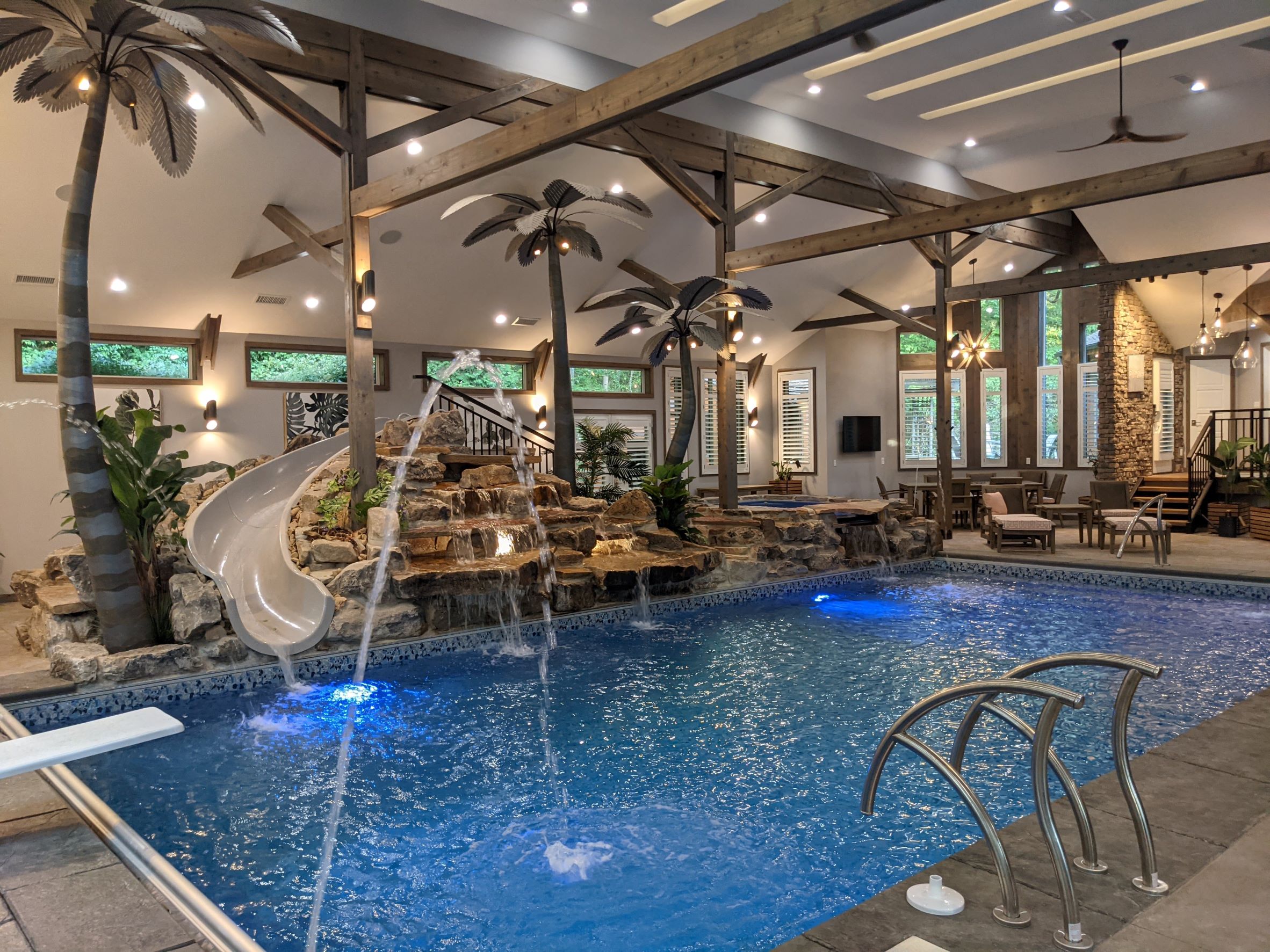 Interior View of Residential Pool Addition to Private Home with Swimming Pool in foreground and stone with waterfalls, hot tub, waterfall in background and palm trees, wood timber beams and columns outside of Pittsburgh Pennsylvania