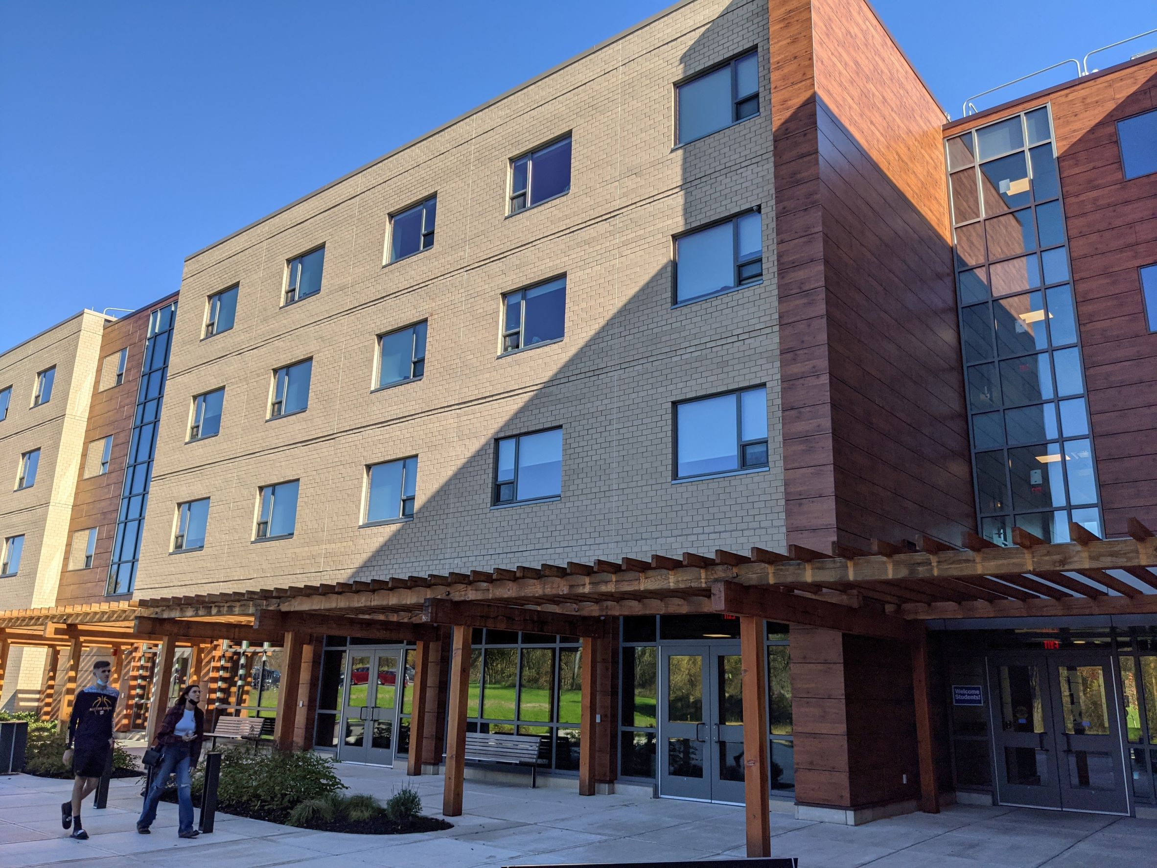 beige brick building with first floor glass and trellis over walkway leading to all glass entry with large wood paneled wall against brick in Central New York State

