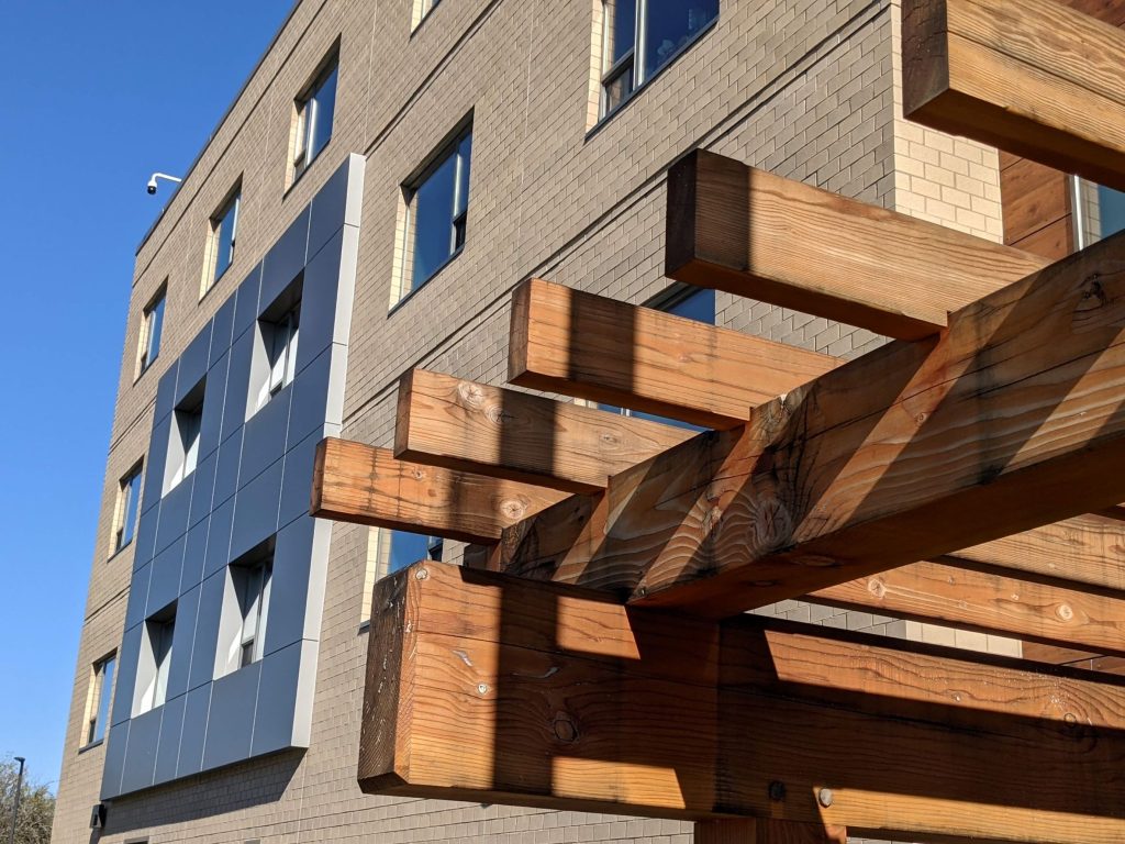 Close up view of wood trellis with beige brick building with silver metallic square projection on dormitory in New York 