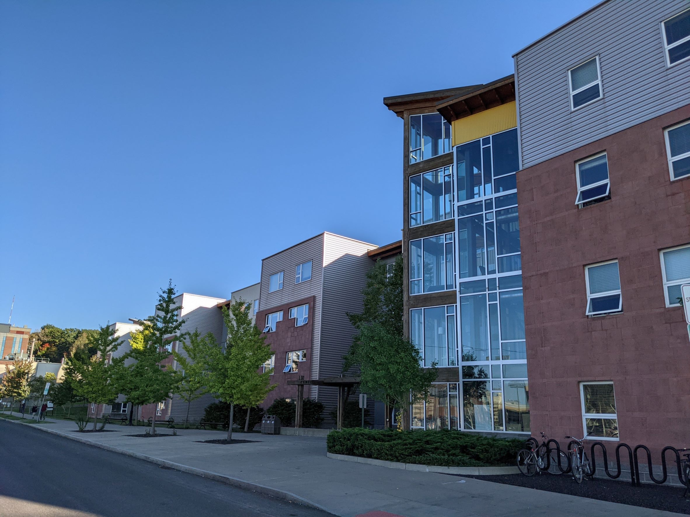 Close up of main entry to student housing building with bright yellow metal panel surrounding glass lounge next to a wood tower that looks like a forest ranger watchtower, surrounded by separate looking brick and metal buildings