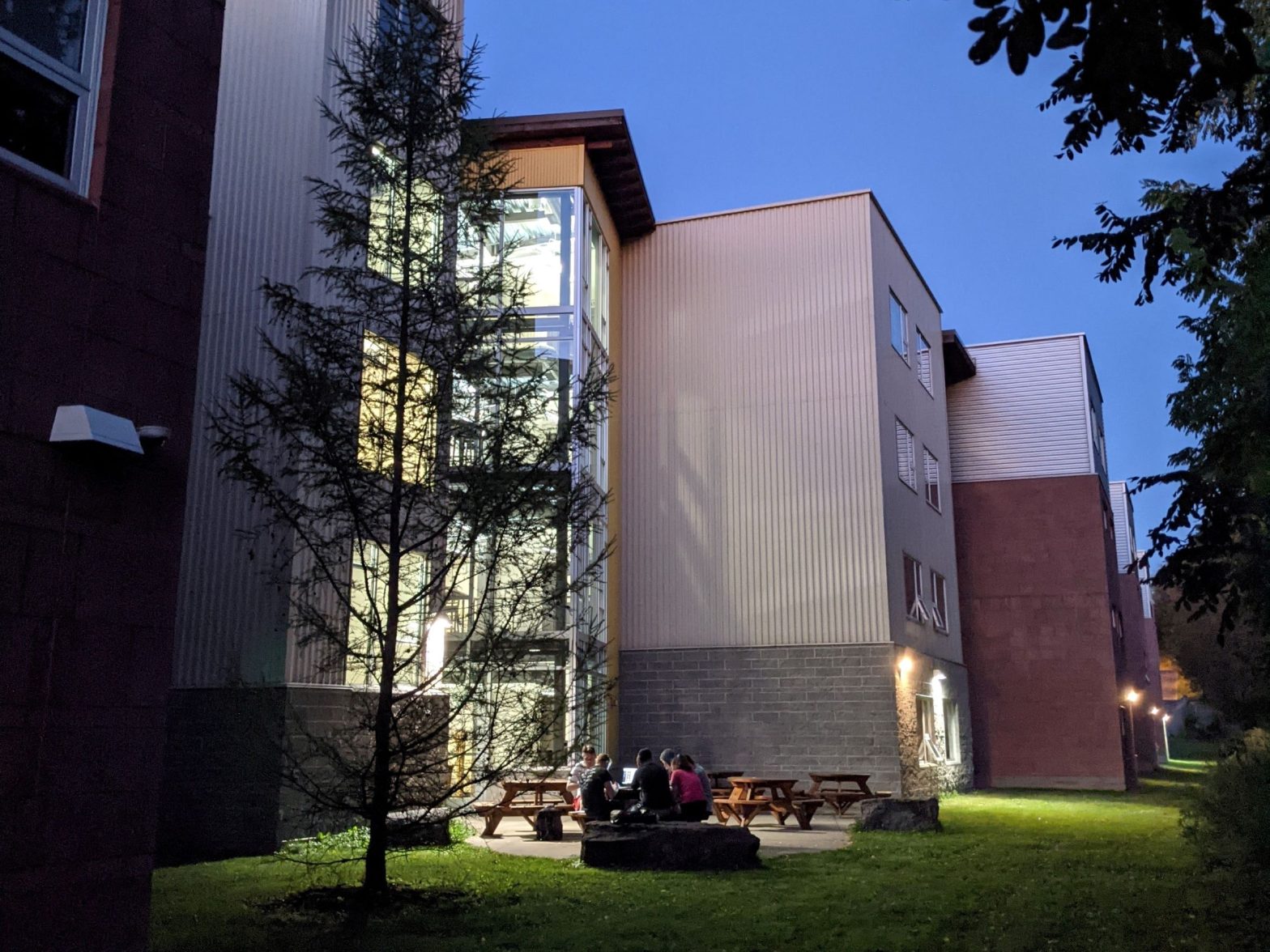 Several students studying on a picnic table next to a boulder and pine tree, against the backdrop of a contemporary gray steel building with a bright yellow and glass tower exuding light at dusk outside of dormitory / residence hall